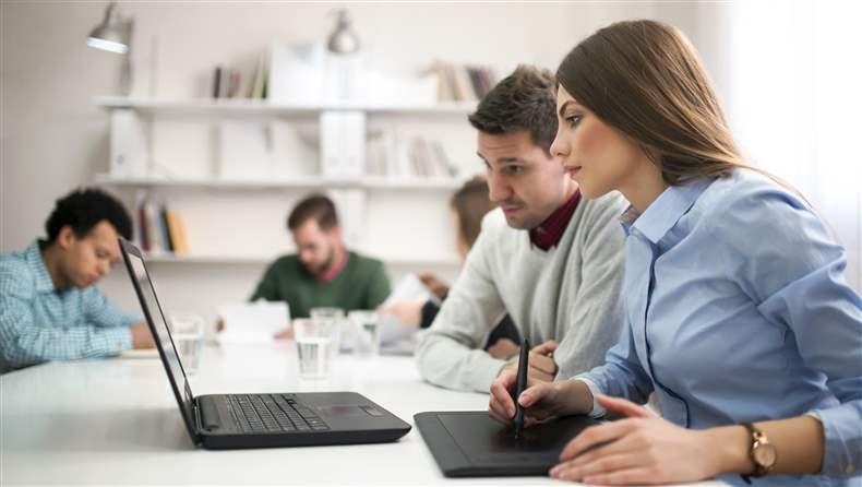 Image Description: Women and man at a table looking at a laptop with other people in the background