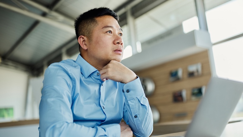 Thoughtful Asian salesman working on laptop in a car shop